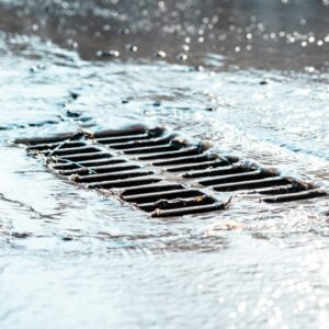 The Grate Of The Storm Sewer After The Rain. The Water Drains Into The Storm Drain. Sun Glare, Defocused Background