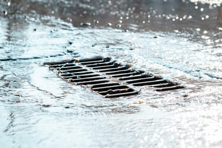 The Grate Of The Storm Sewer After The Rain. The Water Drains Into The Storm Drain. Sun Glare, Defocused Background